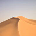 Sand dune under a pale blue sky; warm orange and brown tones.